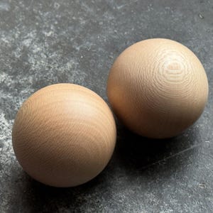 Peut inclure: Deux objets sphériques en bois de couleur brun clair avec un grain de bois visible. Les sphères sont lisses et semblent être faites de bois dur. Les objets reposent sur une surface sombre et texturée.