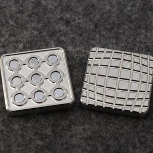 May include: Two square, silver-colored metal fidget toys. One has nine circular magnets arranged in a grid. The other has a grid pattern etched into the surface. Both have a raised border.