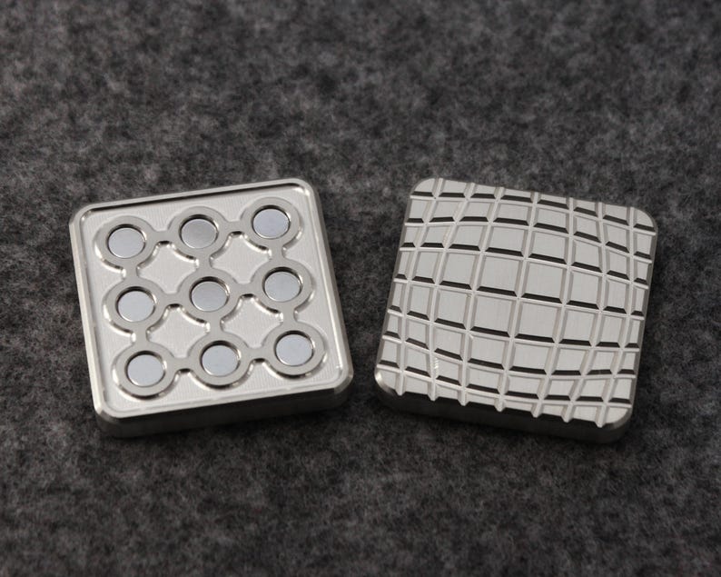May include: Two square, silver-colored metal fidget toys. One has nine circular magnets arranged in a grid. The other has a grid pattern etched into the surface. Both have a raised border.