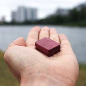 May include: A small, square wooden box with a dark purple stain. The box is sitting in the palm of a hand.