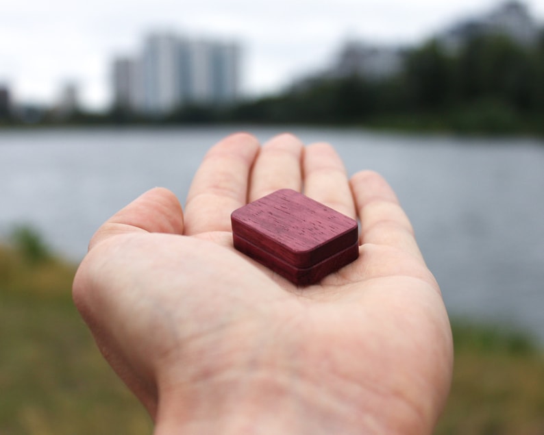 May include: A small, square wooden box with a dark purple stain. The box is sitting in the palm of a hand.