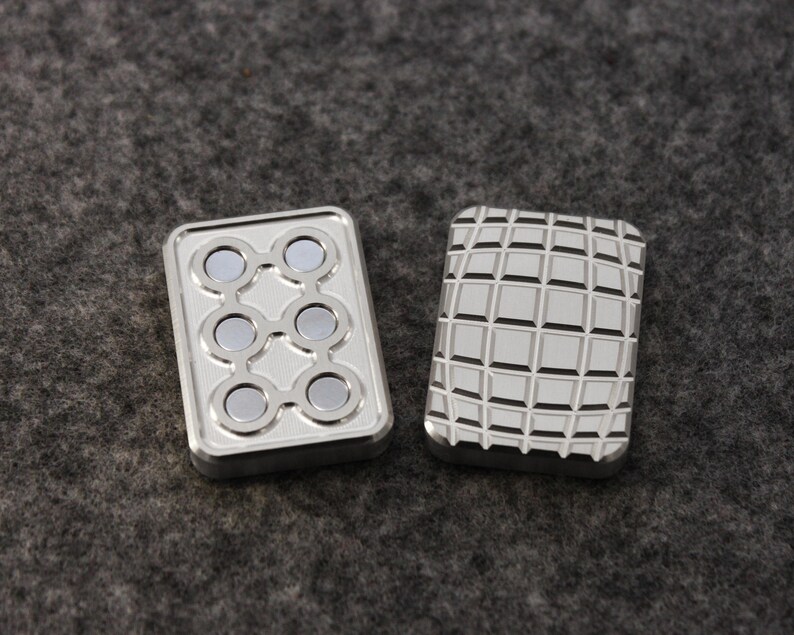 May include: Two silver metal fidget toys. One has six circular magnets arranged in a pattern. The other has a grid pattern. Both are rectangular and have a shiny finish. The background is a textured gray surface.