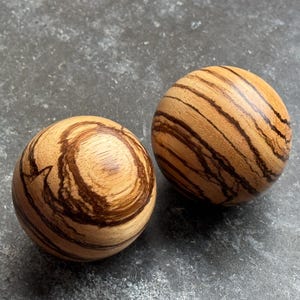 May include: Two wooden spheres with a natural wood grain pattern. The spheres have a light brown base color with dark brown stripes and swirls. The spheres are smooth and round.