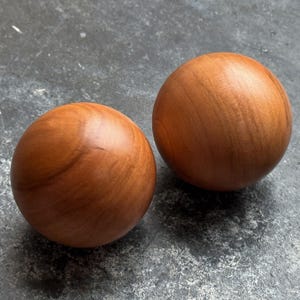 Può includere: Due sfere lisce in legno, con una calda tonalità marrone-rossastra. La finitura lucida rivela le venature naturali del legno. Questi oggetti sferici sono probabilmente decorativi o funzionali, con un diametro di circa 7 cm.