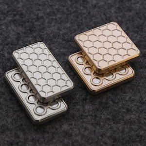 Könnte beinhalten: Zwei Sätze Metall-Fidget-Spinner. Ein Satz ist silberfarben, der andere goldfarben. Jeder Spinner hat eine rechteckige Form mit einem Wabenmuster auf einer Seite und kreisförmigen Vertiefungen auf der anderen Seite. Die Spinner liegen auf einer grauen Oberfläche.