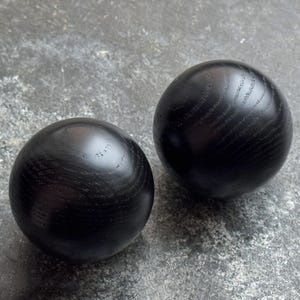 Puede incluir: Dos bolas esféricas de madera maciza negra con un acabado liso y pulido. La veta de la madera es visible en la superficie de las esferas. Las bolas se muestran sobre una superficie gris texturizada.