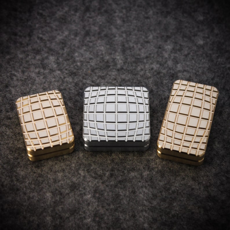 May include: Three metal boxes with a grid pattern. Two are gold-toned, and one is silver. The boxes are rectangular with rounded corners and sit on a dark gray surface. The grid pattern adds a textured look to the boxes.
