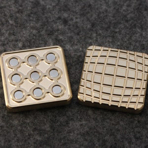 May include: Two square, gold-colored fidget toys. One has nine silver magnets arranged in a grid, the other has a textured, grid-like pattern. The toys are designed to be manipulated and provide a tactile experience.