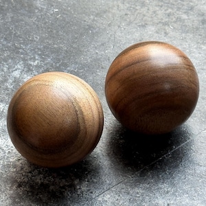 Może przedstawiać: Dwie drewniane kule o wypolerowanym wykończeniu. Kule są w ciepłym brązowym kolorze z widocznymi wzorami słojów drewna. Leżą na szarej powierzchni.