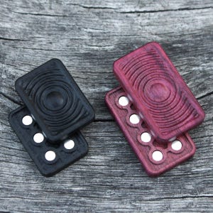 May include: Two wooden pill boxes with a circular design. One box is black and the other is a deep purple color. Both boxes have a row of small, round, white pills inside.