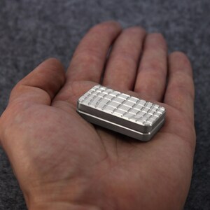 May include: A small, rectangular, silver-colored fidget toy with a textured, grid-like pattern on the top surface. The edges are smooth and the toy is held in an open hand.