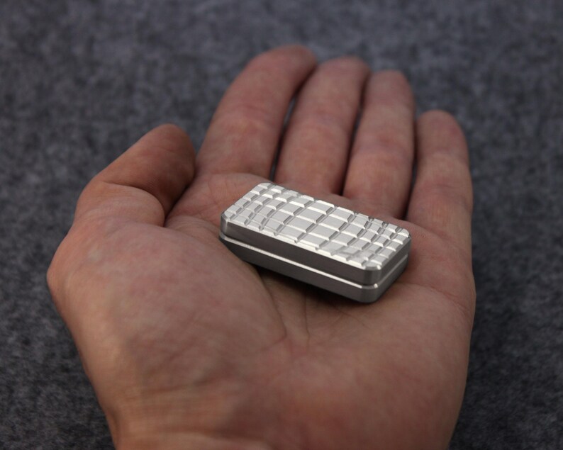 May include: A small, rectangular, silver-colored fidget toy with a textured, grid-like pattern on the top surface. The edges are smooth and the toy is held in an open hand.