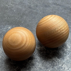 May include: Two spherical wooden objects, light brown in colour, with visible wood grain. The spheres have a smooth, rounded shape and a natural finish. They are displayed on a grey surface.