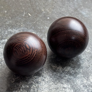 May include: Two dark brown wooden spheres with a polished finish. The wood grain is visible, with lighter brown patterns against the darker background. The spheres are smooth and round, resting on a gray surface.