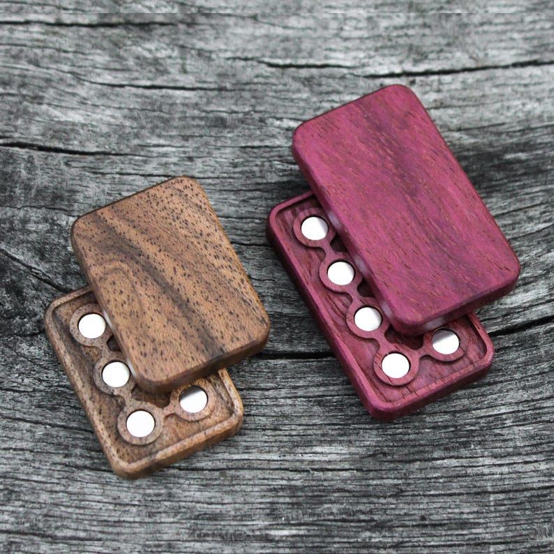 May include: Two wooden boxes with magnetic closures. The box on the left is brown with a natural wood grain finish. The box on the right is a deep purple with a wood grain finish. Both boxes have a circular pattern of eight holes.