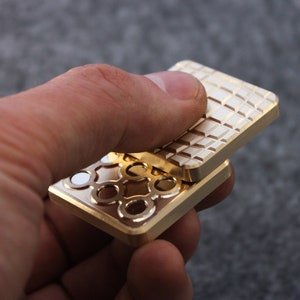 May include: A gold-colored fidget toy with a geometric pattern on one side and a decorative design on the other. The toy is rectangular and appears to be made of metal. The design includes circles and white accents.