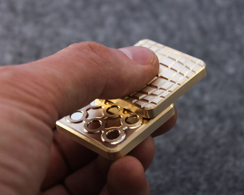 May include: A gold-colored fidget toy with a geometric pattern on one side and a decorative design on the other. The toy is rectangular and appears to be made of metal. The design includes circles and white accents.