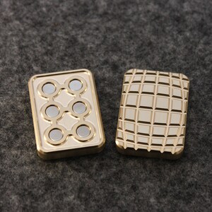 May include: Two rectangular, gold-colored fidget toys. One has six silver magnets arranged in a pattern, the other has a grid-like texture. Both have a raised border and are set against a gray background.