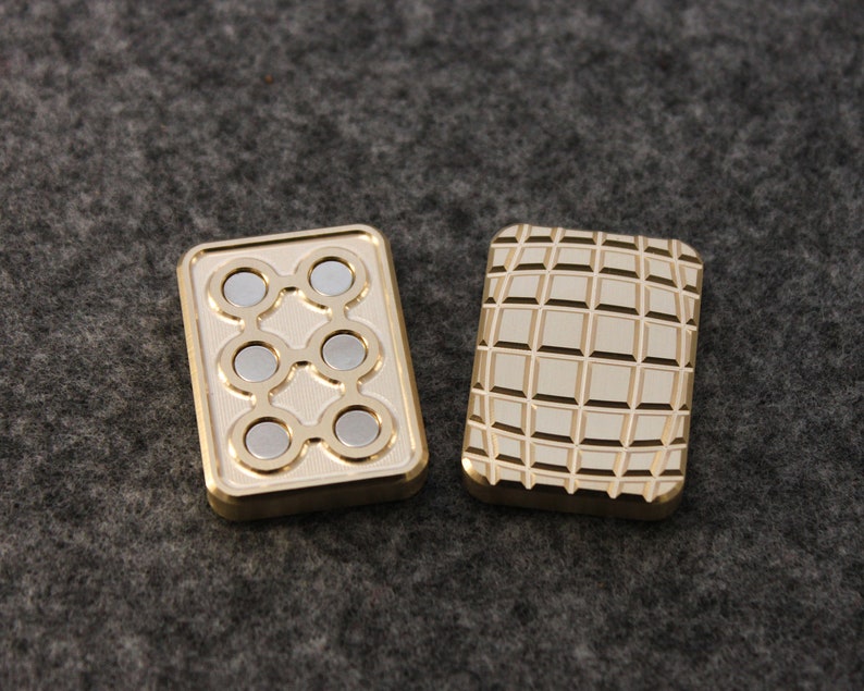 May include: Two rectangular, gold-colored fidget toys. One has six silver magnets arranged in a pattern, the other has a grid-like texture. Both have a raised border and are set against a gray background.