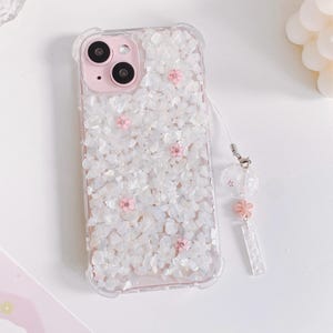 May include: A clear phone case with a white and pink design. The case has a textured surface with small white stones and pink flowers. The case has a clear strap with a pink flower charm.