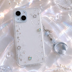 May include: Clear phone case with a white interior, decorated with star and planet charms, and a matching headphone charm. The case has a glittery effect and is designed for a smartphone. The case is decorated with small stars and moons.