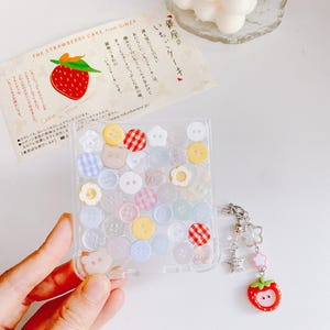 May include: Clear phone case filled with colorful buttons, including floral, gingham, and bear-shaped designs. A strawberry charm with a star and flower dangle hangs from the case. A card with a strawberry illustration is also visible.