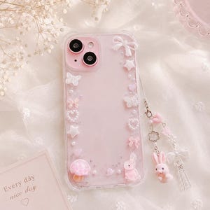 May include: Pink phone case with decorative embellishments including butterflies, stars, bows, and a mushroom. A small pink bunny charm hangs from the case. The case has the text "Every day nice day" on a card.