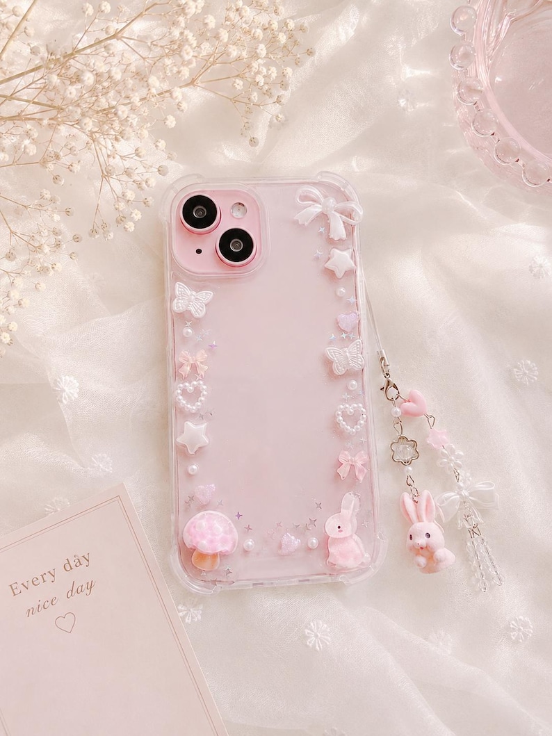 May include: Pink phone case with decorative embellishments including butterflies, stars, bows, and a mushroom. A small pink bunny charm hangs from the case. The case has the text "Every day nice day" on a card.