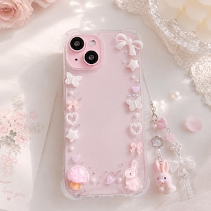 May include: A pink phone case with a clear border, decorated with white bows, stars, butterflies, hearts, and a mushroom. A small pink bunny charm hangs from the case. The case is surrounded by flowers and a card that says "love you".