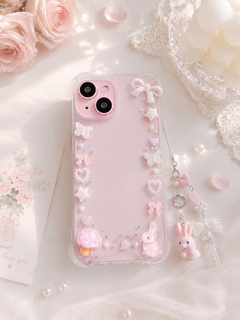 May include: A pink phone case with a clear border, decorated with white bows, stars, butterflies, hearts, and a mushroom. A small pink bunny charm hangs from the case. The case is surrounded by flowers and a card that says "love you".