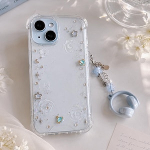 May include: A clear phone case with a light blue background, decorated with stars, moons, and glitter. A blue charm with a headset design hangs from the case. The phone case has a clear border and a dual-lens camera.