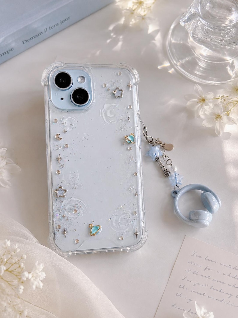 May include: A clear phone case with a light blue background, decorated with stars, moons, and glitter. A blue charm with a headset design hangs from the case. The phone case has a clear border and a dual-lens camera.