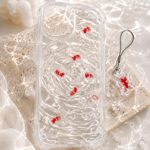 May include: Clear phone case with red goldfish embedded in a water-like design. Includes a matching charm with a goldfish in a bag. The case is on a beige stone surface.