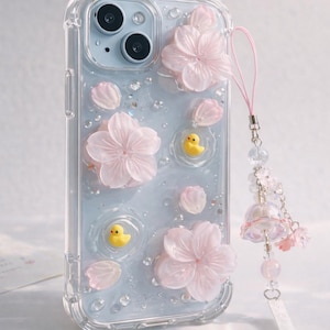 以下が含まれることがあります： 淡い青色の背景に、ピンクの桜の花、花びら、小さな黄色いアヒルのフィギュアが飾られた透明な携帯電話ケース。ピンクのストラップとチャームが付いています。このケースはスマートフォン用に設計されています。