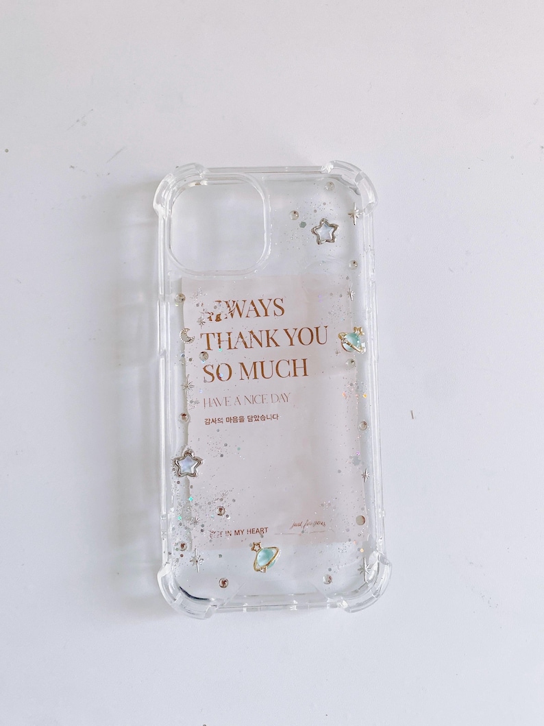May include: Clear phone case with a decorative design. The case features the text "WAYS THANK YOU SO MUCH HAVE A NICE DAY" with star and planet embellishments. The case is made of clear plastic.