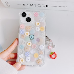 May include: Clear phone case decorated with colorful buttons, bear and flower shapes, and glitter accents. A charm with flowers, stars, and a red strawberry dangles from the case. The phone case is a cute and whimsical accessory.