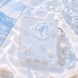 May include: Clear phone case with a blue seal design, starfish, and seashells. The case has a clear strap with a charm featuring a seal, a heart, and a seashell. The case is decorated with a water-like effect.