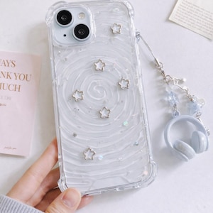 May include: Clear phone case with a white swirl design, silver star accents, and a matching headphone charm. The case has a clear bumper and is designed for a smartphone.