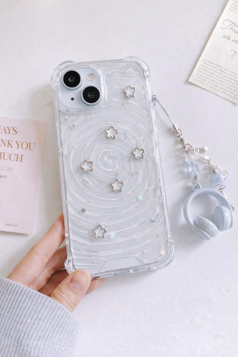 May include: Clear phone case with a white swirl design, silver star accents, and a matching headphone charm. The case has a clear bumper and is designed for a smartphone.