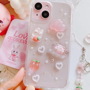 May include: A clear phone case with a pink and white design. The case features a variety of charms, including a pink cloud, a peach, a bunny, and hearts. The case also has a pink and white phone strap.