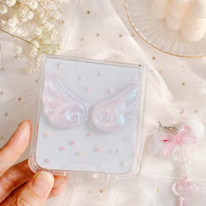 May include: A clear plastic phone case featuring two iridescent, pearlescent angel wings attached to the back. The case is displayed against a light background with small star accents. Beside the case is a small charm with a pink bow and key.