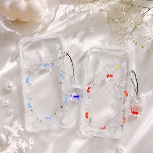 May include: Two clear phone cases with decorative elements. One case features blue accents, the other red. Both cases have small charms attached. The background is a white, silky fabric with decorative elements.