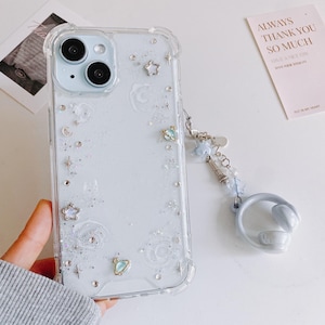 May include: Clear phone case with a celestial design, featuring stars, moons, and glitter. The case has a matching blue wrist strap with star charms and a ring holder. The phone case is held by a person.