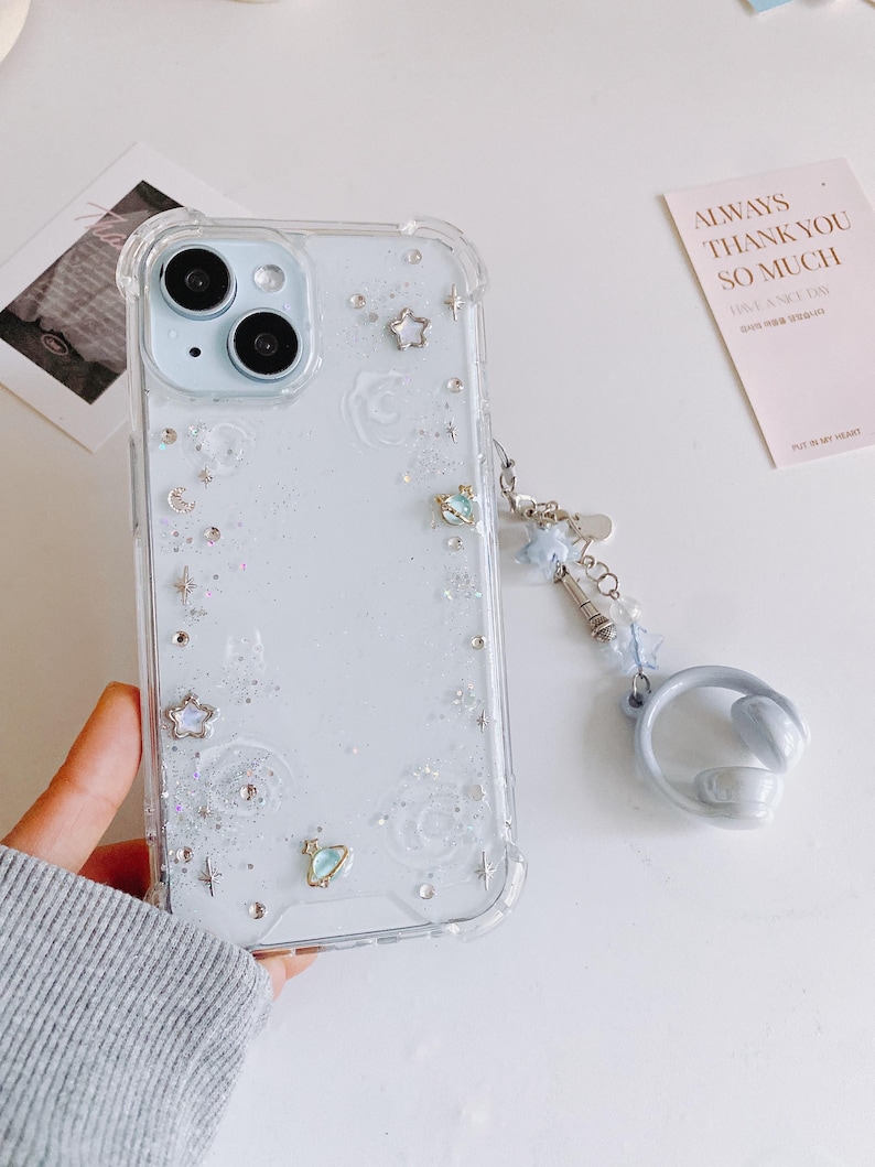 May include: Clear phone case with a celestial design, featuring stars, moons, and glitter. The case has a matching blue wrist strap with star charms and a ring holder. The phone case is held by a person.