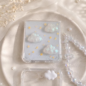 May include: Clear phone case with a light blue background, featuring three white cloud-shaped embellishments, yellow stars, and crescent moons. A second clear case and a beaded strap are also visible.