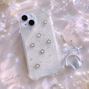 May include: Clear phone case with a white swirl design, decorated with gold star charms. Includes a matching headphone charm and a card that says "Thank you so much". The case is designed for a smartphone.
