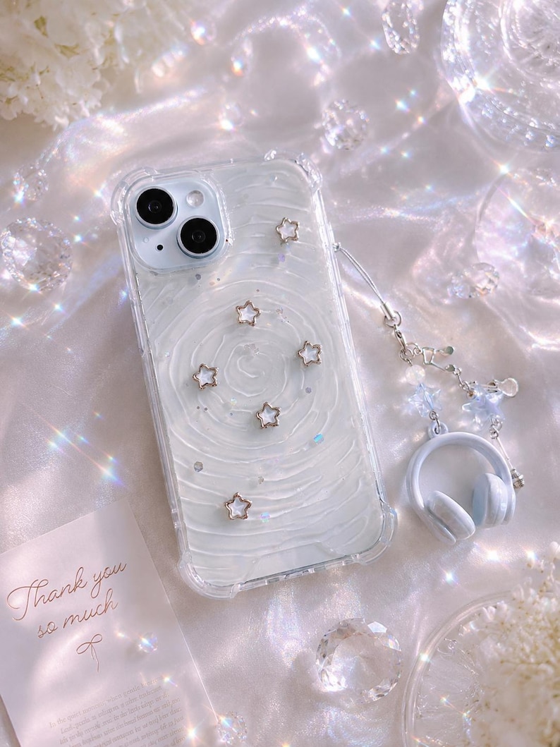 May include: Clear phone case with a white swirl design, adorned with gold star charms. Includes a matching headphone charm and a card that says "Thank you so much". The case is designed for a smartphone.