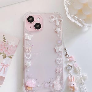 May include: A pink phone case with a clear border, adorned with white bows, stars, butterflies, and a mushroom. A charm with pink hearts, a flower, and a bunny hangs from the case. The phone case is decorated with small pearls.