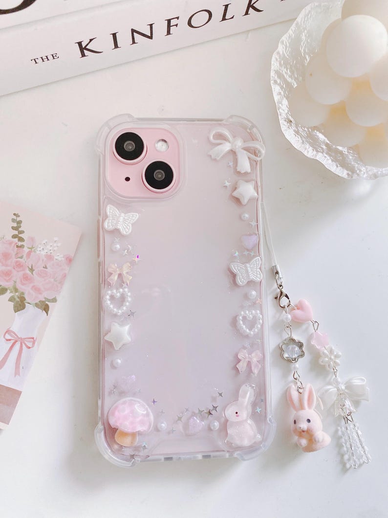May include: A pink phone case with a clear border, adorned with white bows, stars, butterflies, and a mushroom. A charm with pink hearts, a flower, and a bunny hangs from the case. The phone case is decorated with small pearls.