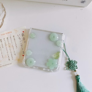 May include: Clear phone case with green floral and circular embellishments. A green tassel and decorative knot are attached. The case is on a white surface, with a book and a small card visible in the background.